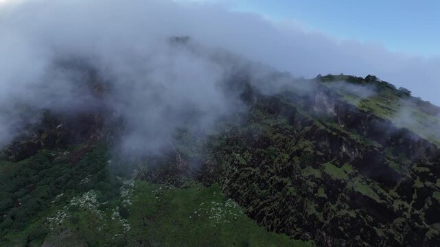 Aerial drone view of a steep, rugged mountain slope covered in green vegetation and enveloped by thick rolling fog and clouds in Indonesia