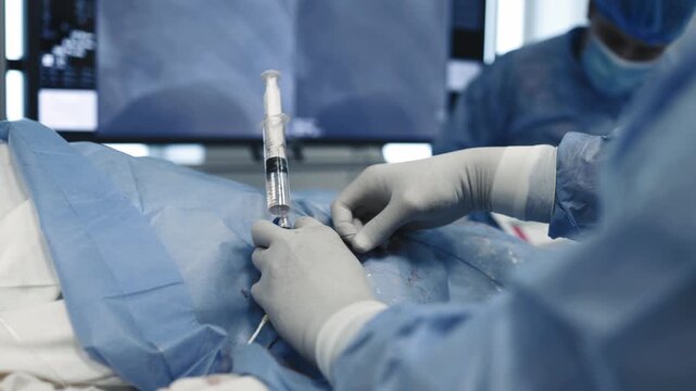 Doctor in gloves working with a syringe and catheter during angiography while vascular imaging is displayed on a monitor.