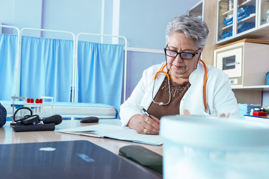 An older female doctor stands in her medical office, looking professional and confident. The portrait reflects her experience and dedication to caring for patients