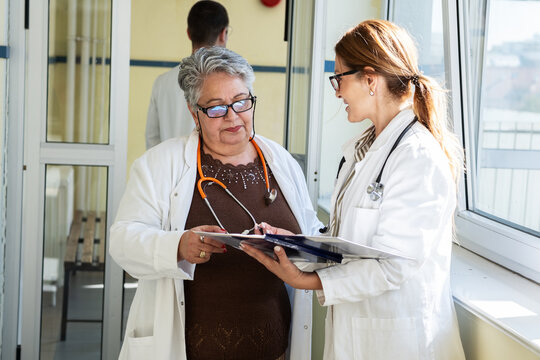 Two female doctors, one senior and one younger, discuss a case in the hallway outside a medical office. They exchange ideas and review information together in a calm and professional setting.