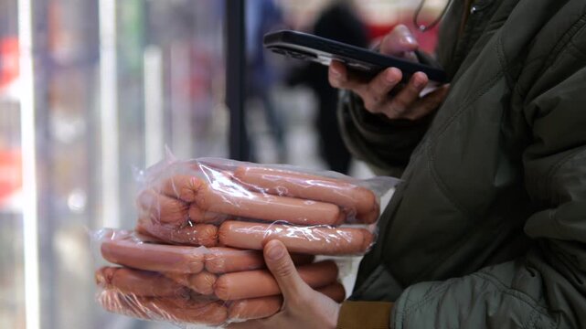 Grocery shopper compares packaged sausages with smartphone in hand inside refrigerated aisle. Customer checks groceries while choosing processed meat, making practical food purchase in supermarket.