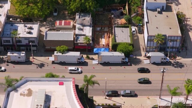 Film production trucks parked in a row behind orange traffic cones on a Los Angeles street. Aerial view of passing cars, palm trees and a wooden frame construction site on a sunny summer day.