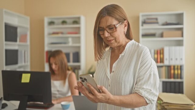 Woman holding smartphone and reading messages while standing by a desk with computer and colleague at workstation in building; focused collaboration.