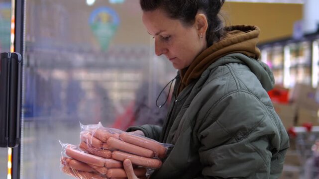 Woman studies packaged sausages during grocery shopping, calmly reviewing her meat selection near chilled cases. Shopper holds several transparent packs and checks contents before making choice.