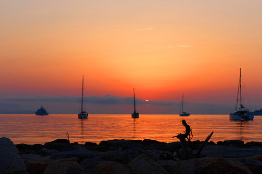 Sailboats and yachts silhouetted at sunset over calm Mediterranean waters with rocky shore, Antibes.