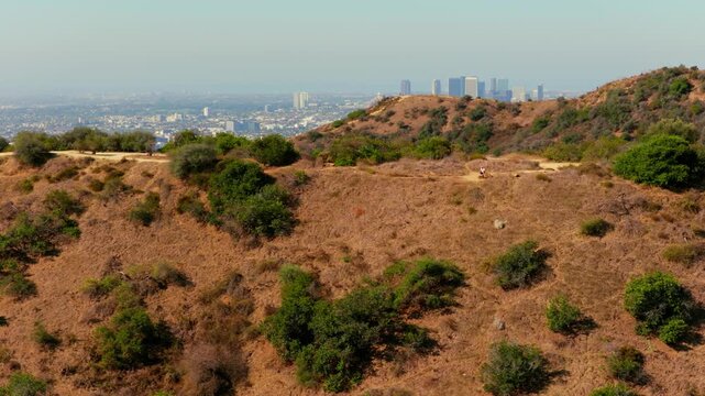 A runner on a dirt trail crosses dry golden grass and green shrubs in the Los Angeles hills. Century City towers and the hazy city sprawl stretch to the horizon. Sunny summer aerial of chaparral.