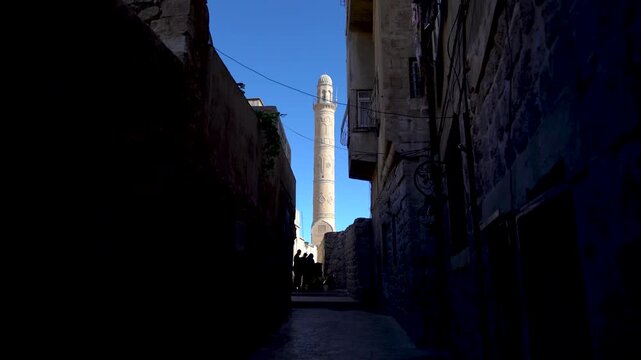 Ulu Cami minaret of the Great Mosque of Mardin, Turkey, framed by a dark narrow street of the old town, with silhouettes of people at its end