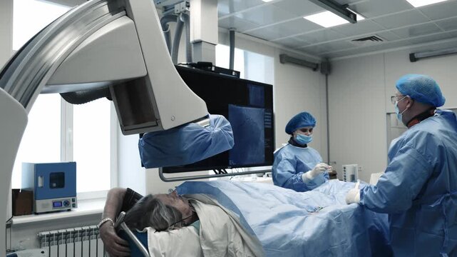 Doctor working beside a patient under a C-arm angiography system during a catheter procedure in a hospital cath lab.