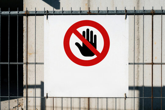 Red, white stop sign attached to metal wire fence at construction site. No entry warning symbol with black hand icon prevents access to danger zone. Safety notice protects people from hazards in