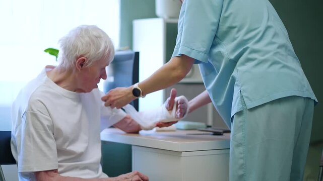 Medical staff gently bandaging frail senior citizen with broken arm in clinical treatment room. Compassionate care highlights osteoporosis treatment and bone health for vulnerable elderly individuals