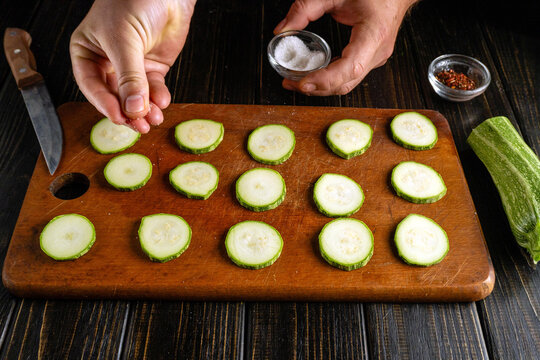 Hands season sliced zucchini with salt above wooden cutting board. Fresh vegetable rounds are arranged beside kitchen knife and bowl of red pepper flakes for simple meal preparation.