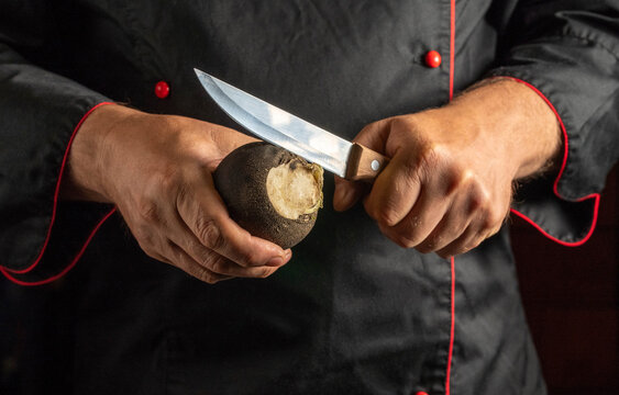 Chef cuts into black radish with steel kitchen knife, revealing its light center. Cook holds round root firmly, focused on ingredient preparation in dark apron with red edging, as blade trims its