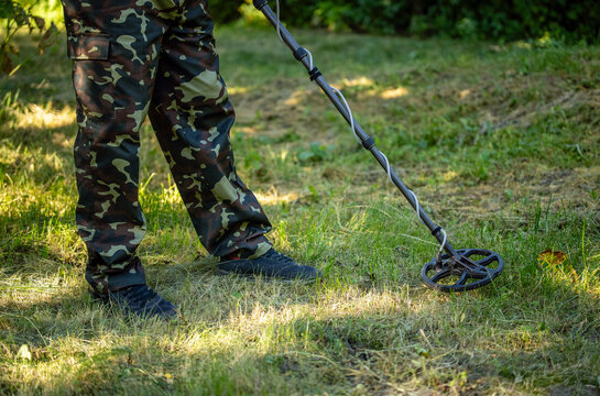 Person in camouflage trousers sweeps metal detector coil over grassy ground. Detector operator searches outdoors in woodland light, holding equipment close to earth while wearing sturdy boots.