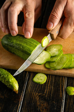 Cook cuts cucumber into thin rounds on wooden board, holding vegetable steady beside sharp knife. Crisp slices gather nearby, suggesting fresh ingredient preparation for home cooking.