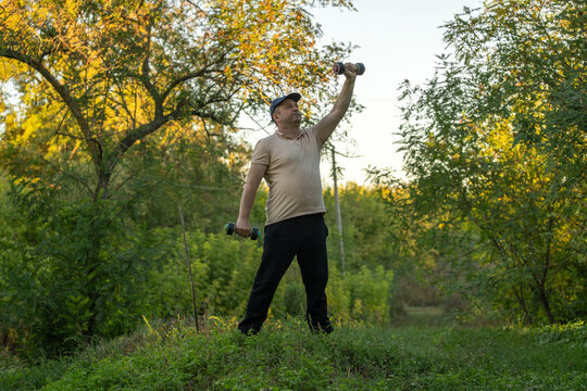 Middle aged man lifts dumbbell overhead while holding another weight at his side, exercising outdoors. Focused and steady, fitness enthusiast stands on grassy woodland path beneath autumn trees.