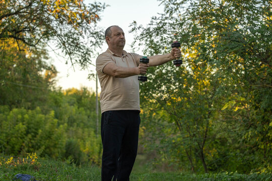 Focused middle aged man performs standing dumbbell exercise in leafy park, holding one weight in each outstretched hand. Outdoor fitness gives his workout calm, natural setting, with arms extended