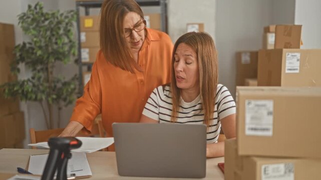 Woman points to laptop while partner rubs face and sorts cardboard parcels at small business desk in building; frustration mentoring.