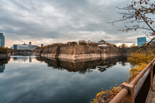 View of Osaka Castle's moat and stone walls with city skyline under cloudy blue sky in autumn season