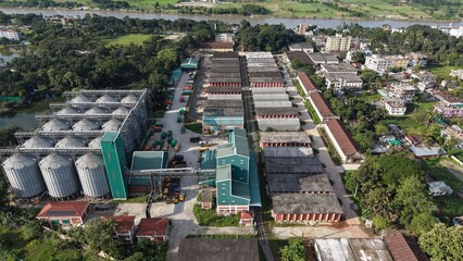 Aerial view of a large industrial grain silo complex with storage warehouses and transport trucks located near a river in a rural landscape © ridwanhassan