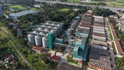 Aerial view of a large industrial grain storage facility with tall metal silos and warehouse buildings located near a river and green landscape © ridwanhassan
