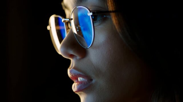 Woman in round eyeglasses keeps her gaze forward in side profile as camera moves closer, blue reflections filling lenses while warm edge light shapes her face against dark background