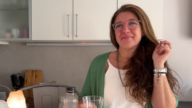 Content mature woman with eyeglasses relaxing in her kitchen. She is smiling and enjoying the moment as she drinks water.