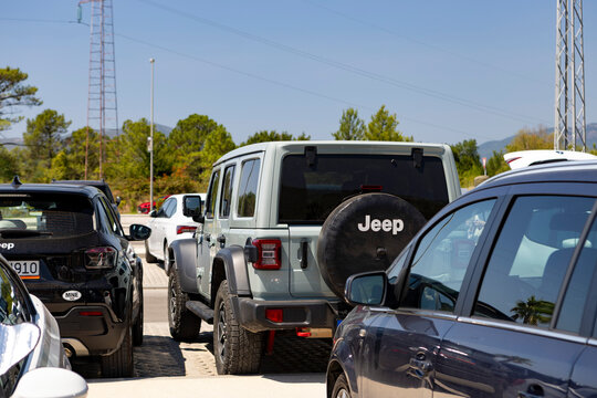 August 3, 2026, Kotor, Montenegro, Sunny outdoor parking lot with rows of parked cars beneath palm trees and paved islands, with distant mountains and clear blue sky in daytime
