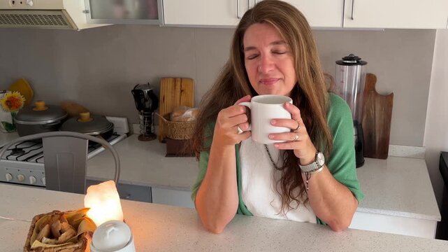 Calm and relaxed woman enjoying a serene moment in her kitchen while drinking a hot beverage from a white mug