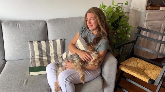 A woman of serene aspect is seated on a sofa in a modern living room, relaxed as she pets her charming dog