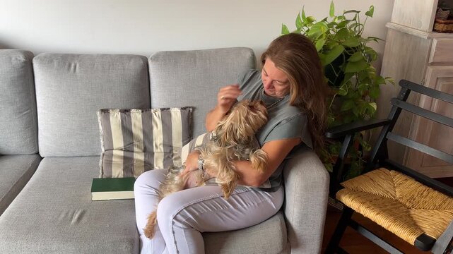 A serene, mature woman sits on a sofa in a modern living room, smiling as she pets her cute dog. A concept of calmness and peace of mind.