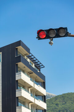 Red traffic light on streetlight pole above modern apartment building with balconies under clear blue sky and green hillside background