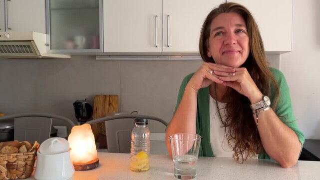 A middle-aged woman is sitting pensively in her kitchen, relishing a calm and happy moment.