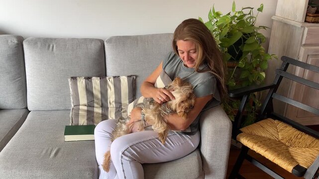 A middle-aged woman is lounging on a sofa in a contemporary living room, looking happy as she strokes her cute dog.