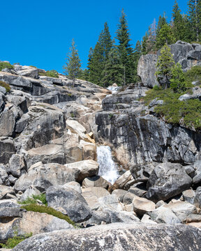 Bassi Falls, Eldorado National Forest. Large boulders and patches of green can be seen nearby. Tall trees rise in the background under a clear blue sky.