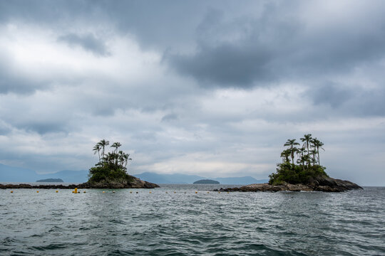 Botinas Islands from a distance in the ocean of Angra dos Reis. Palm trees grow on the islands. The sky is cloudy.