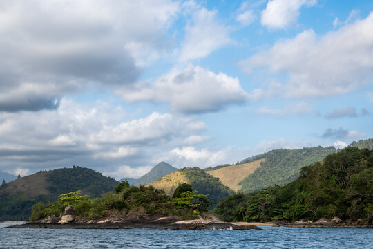 A small island in Angra dos Reis  with green trees and hills in the background. The sky has clouds and looks bright. It is a daytime setting.