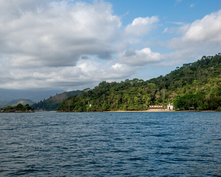 The ocean and green Atlantic forest at Angra dos Reis. Ruins of buildings are visible along the shore, set against a backdrop of hills and cloudy skies.