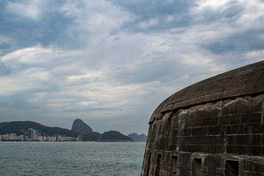 Copacabana Fort stands at the edge of the water in Rio de Janeiro. In the background, mountains rise and buildings dot the shoreline under a cloudy sky.