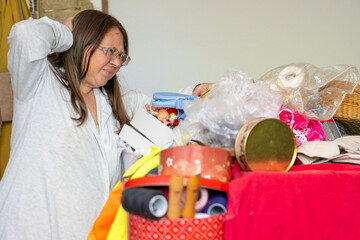 Stressed Middle aged woman with glasses raises hands in frustration standing before pile cluttered items, Home organization sorting household items, Cleaning routine clutter lifestyle transformation © kittyfly