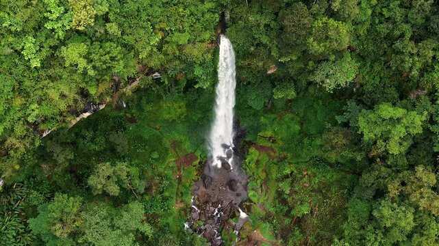 Aerial view of Waterfall Kayu Aro cascading down through dense tropical rainforest with lush green foliage and rocky terrain in Kayu Aro, Jambi, Indonesia.
