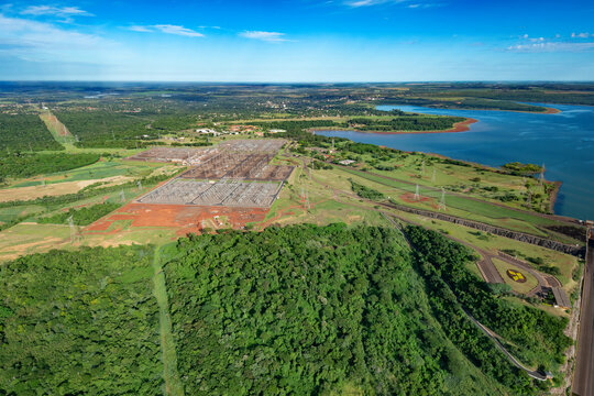Aerial view of an electrical substation beside the Itaipu Dam in Brazil, with rows of switchyard equipment, transmission pylons and power lines surrounded by forest and the blue reservoir.