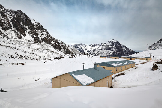 Snow surrounds mountain refuge buildings at Portillo in the Chilean Andes, where low structures with metal roofs and chimneys sit beneath steep, rocky high-altitude peaks.