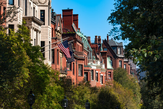 Traditional brick brownstone buildings along Commonwealth Ave in the historic Back Bay neighborhood of Boston with an American flag flying among green trees.