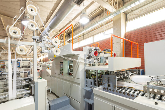 A folder gluer machinery system in operation at a printing plant for the creation of paper bags with paper rolls feed mechanism and automated rollers in a modern industrial factory setting.