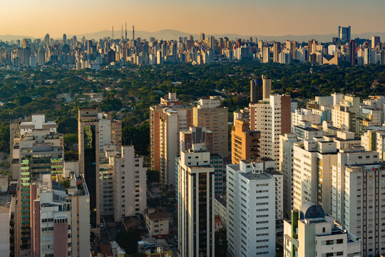 A wide panoramic view over the cityscape of the city of S&atilde;o Paulo in Brazil showing dense high rise buildings and lush green urban trees under a warm golden sunset glow.