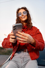 Naklejka na ściany i meble A smiling young woman in a red jacket holding a smartphone outdoors. Concept for technology, lifestyle, and social media communication in modern days.
