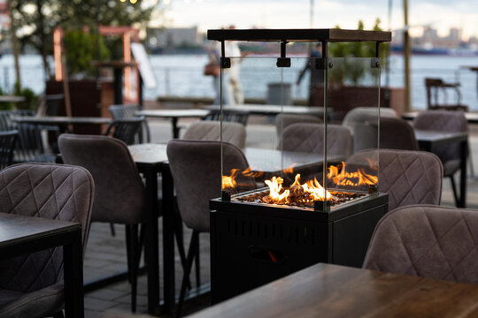 Gas fireplace burns inside glass enclosure among empty restaurant tables near waterfront. Outdoor heater warms vacant terrace during cold autumn off season.