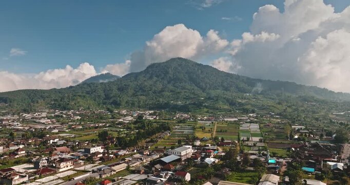 Bali village nestled in a tropical landscape, featuring houses, road, agricultural fields and farms at the foot of lush green mountains under a blue sky with white clouds. Aerial view drone flight