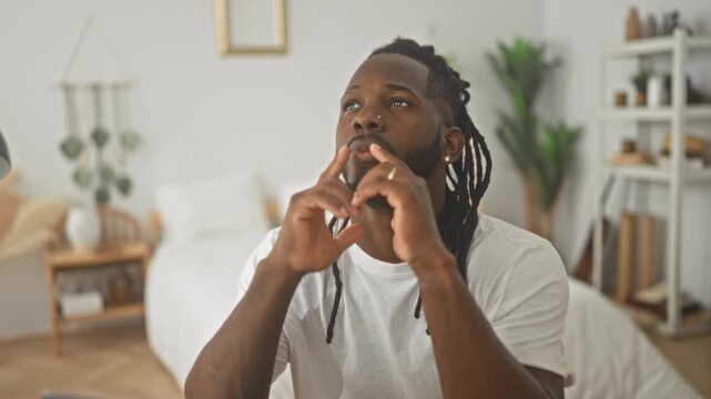 Young african american man clasping hands near mouth at a desk with laptop in a sunlit studio bedroom, bed and shelving visible; contemplation focus.