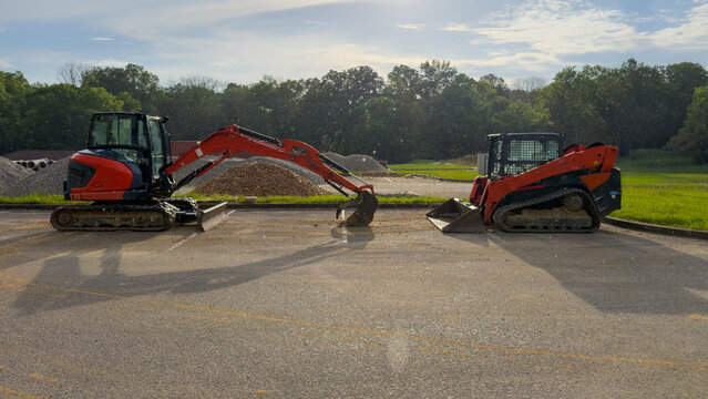 Red compact excavator and tracked skid steer loader face each other on an asphalt parking lot, with gravel piles, grass, and trees behind under a dramatic blue sky